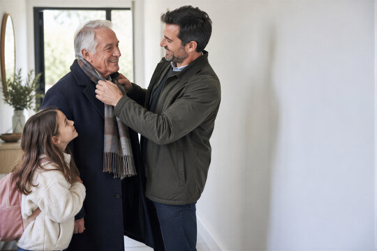 Caring adult son helping his senior father with a scarf in the hallway while a young girl looks on