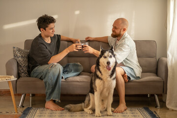 Father and teenage son sit on sofa while husky waits in front of them, generation difference meaning nothing here, scene about being close. Wide shot. Concept of generation talk of father and son © iwavephoto