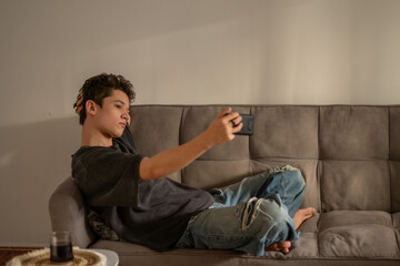 Fototapeta premium Teenager lies on sofa holding phone above himself for new shot, blogging repeated until frame feels right, emotion that stops scrolling. Wide shot. Concept of blogging habits of teenage boy