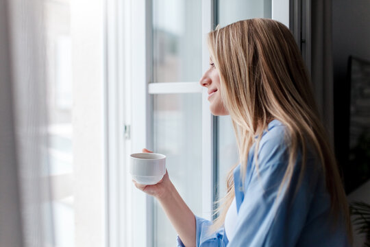 Young woman enjoying coffee, morning routine, calming drink in hotel. Happy girl relaxing by opened window. Slow travel, mindfulness, female mental health. Solo traveler in vacation. Airing out room