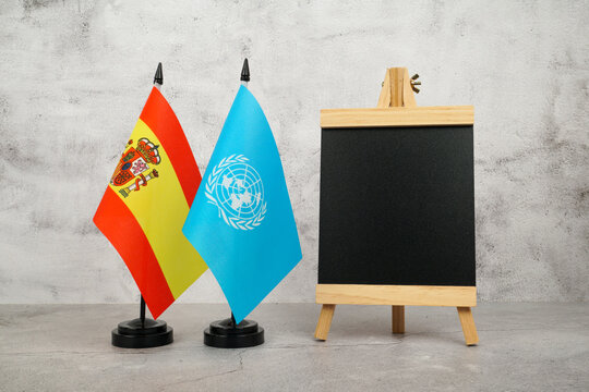 Spain and United Nations flags beside a blank chalkboard on a neutral background with copy space.