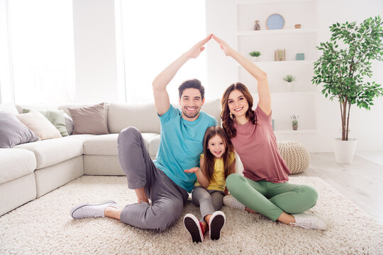 Happy young family at home sitting in a bright living room forming a roof with their arms to symbolize house, togetherness and love while smiling and relaxed on a cozy carpet