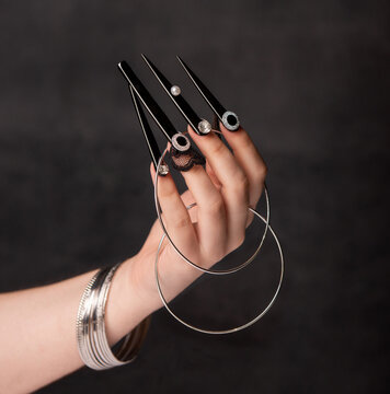 Hand displaying ornate black elongated nails with metallic rings and bracelets against dark background
