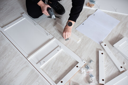 Person assembling flat pack furniture with power drill on wooden floor indoors