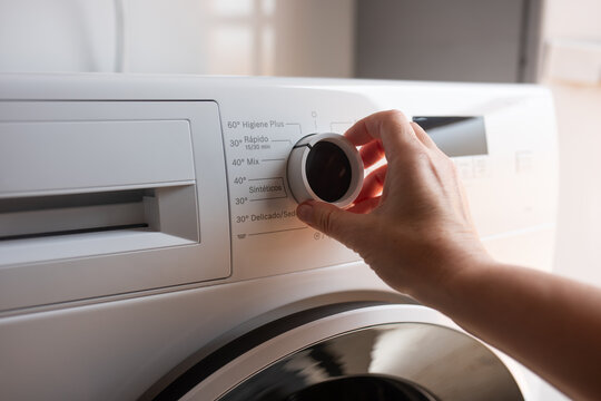 Hand adjusting washing machine cycle dial on modern white laundry appliance indoors