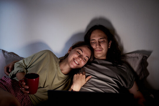 Young adult Caucasian couple watching movie on laptop in bed together at night. Image useful for streaming service advertising, entertainment apps, digital media campaigns