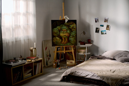 Artist bedroom studio showcasing oil painting on wooden easel beside unmade bed and vinyl turntable. Suitable for creative lifestyle branding and interior design promotion