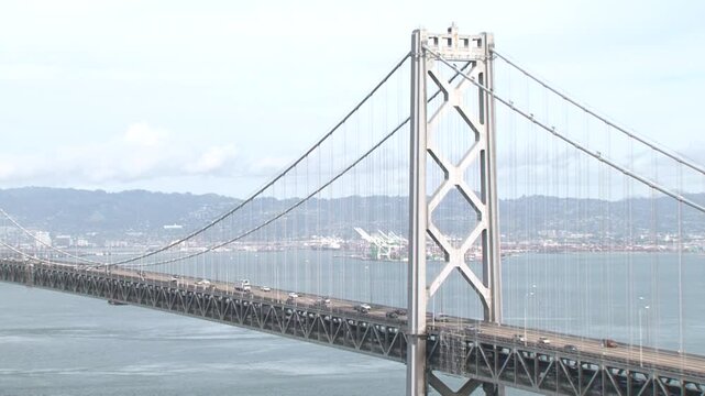 This 2015 video frame features a wide view of a Bay Bridge suspension tower. Cables drape down to the deck where traffic flows, set against the distant Oakland port and rolling hills.