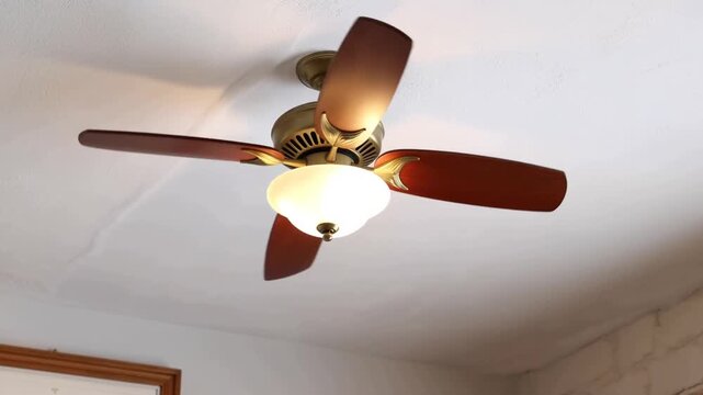 Slow motion tracking shot of ceiling fan blades rotating above bedroom ceiling with warm glowing light fixture