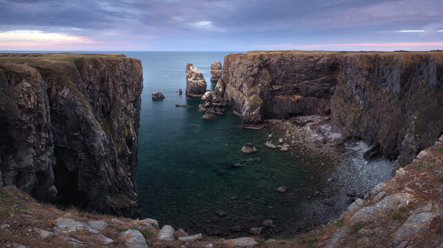 Dramatic sea stacks rise between rugged coastal cliffs at Elegug Stacks, Wales, under soft dawn light above calm dark water.