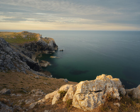 Rugged limestone cliffs and rocky headland overlooking calm coastal waters in Wales, with warm evening light illuminating the foreground rocks.