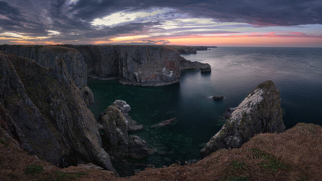 Dramatic sea stacks rise between rugged coastal cliffs at Elegug Stacks, Wales, under soft dawn light above calm dark water.