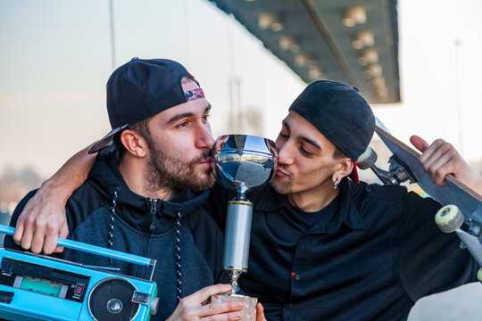 Two young male skateboarders celebrating competition victory, sharing a victorious kiss with a trophy