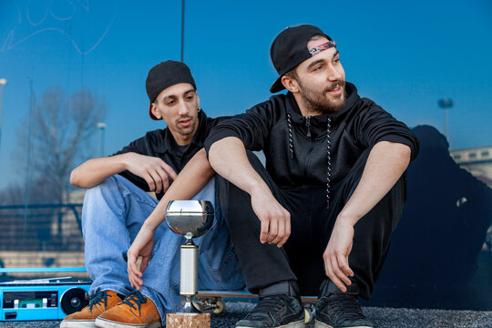 Two young men sitting outdoors with a trophy, skateboard, and boombox, celebrating a win