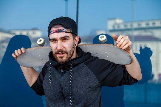 Young man with a beard holding a skateboard on his shoulders, standing in an urban setting