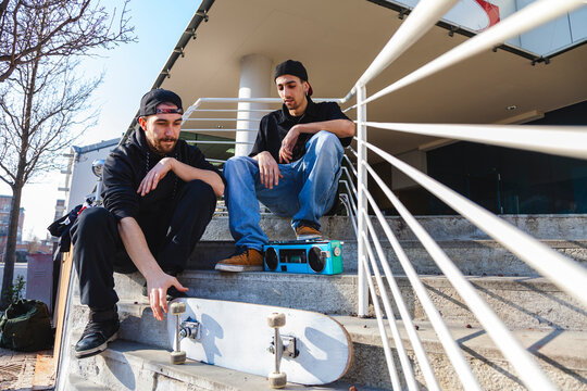 Two friends relaxing on city steps with a skateboard and boombox, embracing urban youth culture