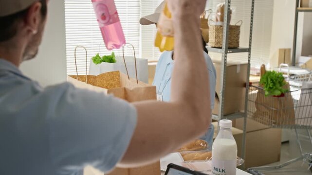 Caucasian worker checking online order form on tablet while Hispanic woman packing fresh groceries into paper bags. Ideal for e-commerce delivery logistics and retail food supply marketing