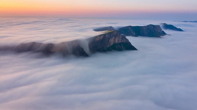 Aerial tracking shot of mountains with cloud cascade flowing over peaks above sea of clouds at sunrise