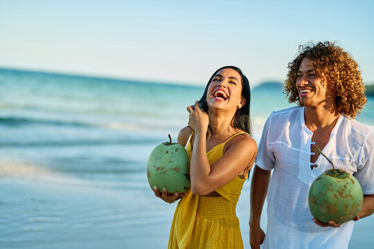 A smiling couple strolls barefoot along a tropical beach, each holding a fresh coconut as gentle waves lap the shore. The woman wears a yellow dress and the man white linen clothing, creating a relaxe