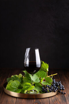 Elegant glass of dark red wine with fresh green and dark grapes and leaves on a brass tray presented on a wooden table with a black textured background.