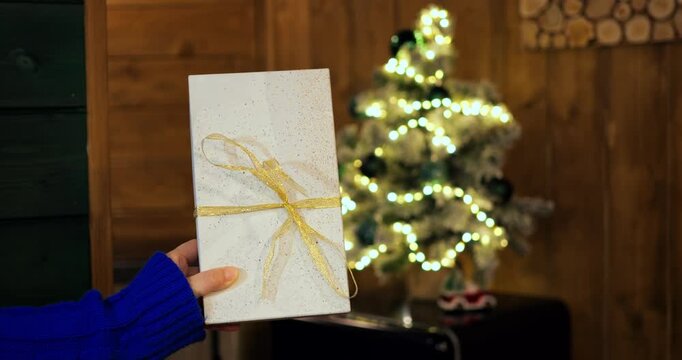 Person wearing blue knit sweater holds wrapped gift box tied with gold ribbon in cozy room. Festive holiday present shines near glowing Christmas tree, bringing joyful celebratory spirit.