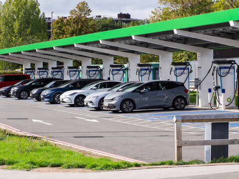 Row of electric cars charging at outdoor station with green canopy, visible charging cables and plugged vehicles beside trees and parking lot pavement