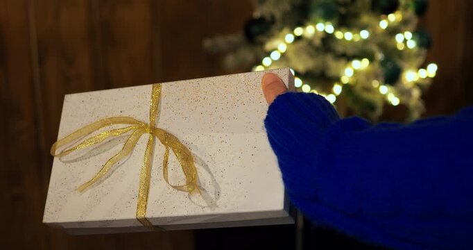 Hand holds elegant white gift box tied with shimmering gold bow during winter celebration. Glowing lights on blurred Christmas tree create warm holiday mood and joyful atmosphere indoors.
