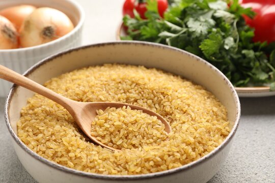 Raw bulgur in bowl, spoon and other products on table, closeup