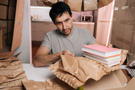 Young man packing books with brown paper into cardboard boxes, preparing orders for shipment from small home office setup, managing inventory and fulfilling customer deliveries.