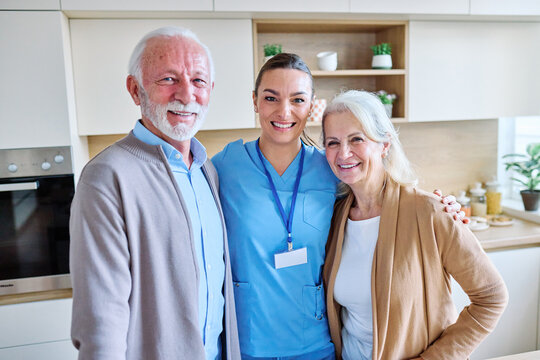 Senior couple smiles warmly with a caring nurse. They stand together in a cozy, modern kitchen. The nurse's professional attire conveys trust and support. Natural light fills the space