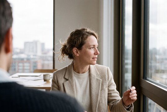 Confident business woman in blazer discussing ideas during an office meeting by a large window overlooking the city