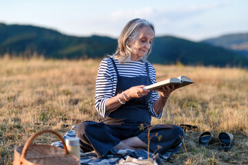 Fototapeta premium Older woman enjoying peaceful solo picnic and reading in countryside.