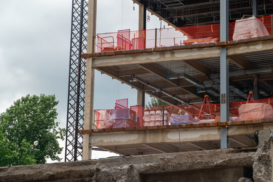 Multi-story building under construction featuring exposed concrete and steel framework, orange safety netting, and building materials against a cloudy sky, representing urban development and growth.