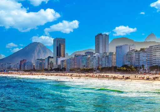 Stunning view of Ipanema beach in Rio de Janeiro, Brazil. Clear blue ocean, sandy coast filled with people, city skyline, and majestic mountains under a sunny sky.