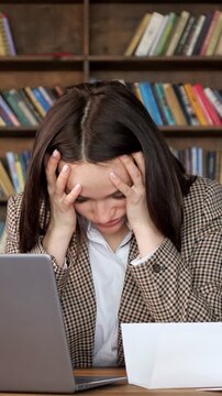 Frustrated brunette businesswoman feeling stressed and tired from overwork, experiencing a severe headache and burnout while sitting in a library, unable to cope with her professional problems