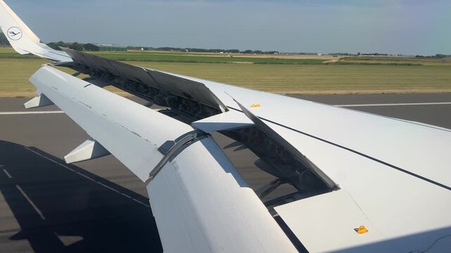 Close-up view of an airplane wing with deployed spoilers during landing, showing aerodynamic control surfaces, runway and airport landscape on a clear sunny day.