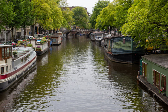 Canal in Amsterdam featuring moored houseboats and historic brick bridge framed by lush green trees in the Netherlands