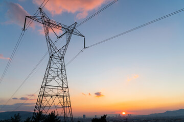 High voltage electricity pylon stands against a colorful sunset sky with power transmission lines stretching across the horizon above a distant city skyline © bulentbaris