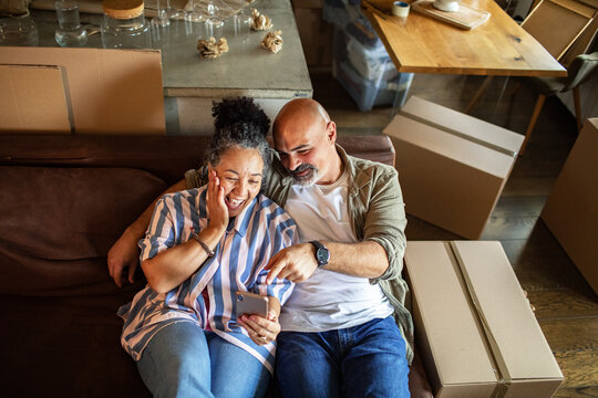 Happy mature couple checking smartphone in new home with moving boxes