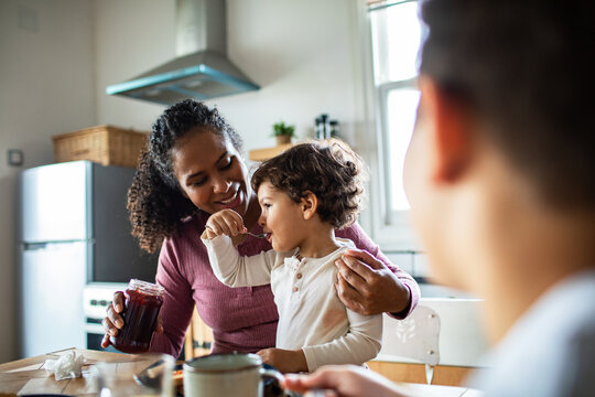 Mother helping toddler taste jam in home kitchen