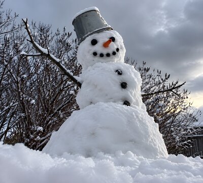 Lopsided snowman in a drifted backyard with coal eyes twig arms and an upturned tin bucket hat