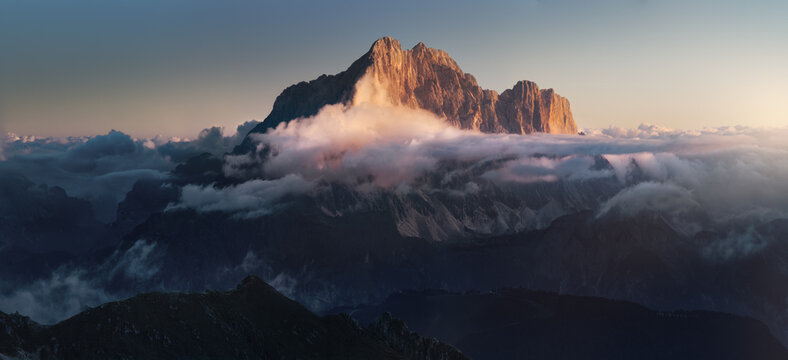 Monte Civetta rises above drifting clouds with its massive rock walls illuminated by warm sunset alpenglow in the Italian Dolomites.