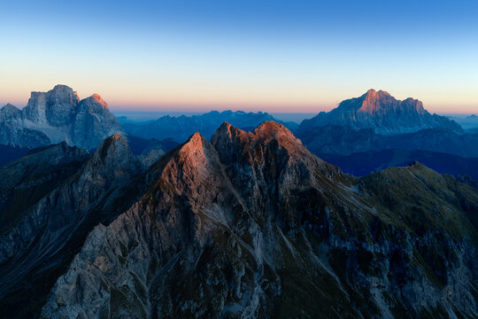 Aerial sunset view across the rugged Croda da Lago ridge and Cima Ambrizzola toward Sorapiss and Antelao, with warm alpenglow illuminating the Italian Dolomites.