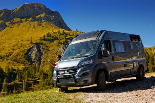 Active woman traveler beside a camper van in a scenic autumn alpine landscape with golden mountain slopes, larch trees and rugged peaks in the Italian Dolomites.