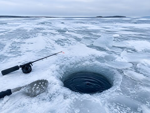 Round fishing hole cut through pale lake ice with dark water at rim beside short rod and scoop