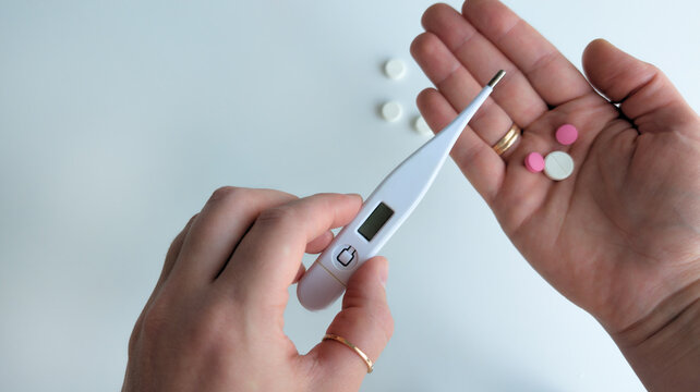 Top view of female hands holding digital thermometer and group of medical pills for cold or fever treatment