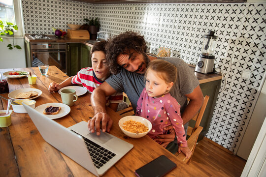 Father helping children with laptop during breakfast in home kitchen
