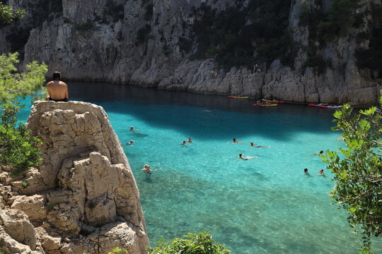 Calanque d'En-Vau, Cassis, France - September 7th 2026: people swimming and kayaking in turquoise waters in Mediterranean sea cove while man sits on rock on a height