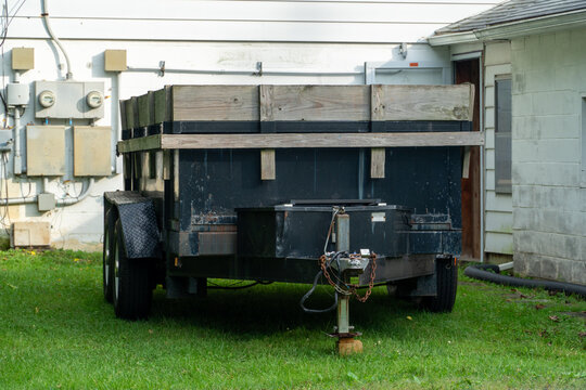 Old, rusty, and weathered black utility trailer with wooden side extensions parked on green grass next to a white residential building with external electrical meters, showing wear and tear.