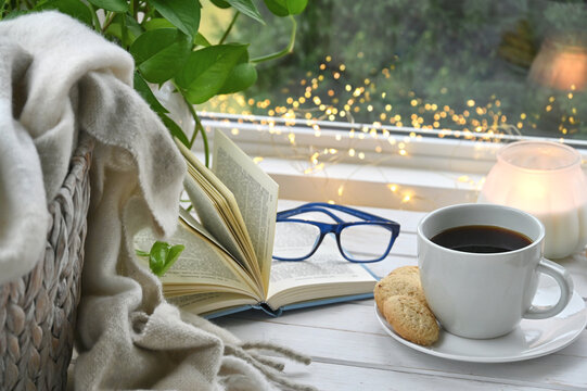 Cozy interior tea coffee mug by window with book, blanket, lights
